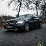 En slank sort Mercedes C300 Coupe parkerer på en våd gade og praler med sit luksuriøse design på baggrund af nøgne efterårstræer og en dyster himmel, der emmer af elegance og kraft.