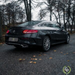 En elegant sort Mercedes C300 Coupe parkeret på en fugtig vej, omgivet af nedfaldne blade, signalerer en humørfyldt efterårsstemning med overskyet himmel.