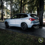 En elegant BMW X3 xDrive30e hybrid SUV med sporty sorte fælge er parkeret langs en skyggefuld kørebane, omgivet af frodige grønne træer, der viser en blanding af luksus og natur.