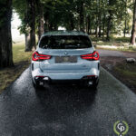 En sølvfarvet BMW X3 xDrive30e plug-in hybrid parkeret på en våd vej foret med træer, med karakteristiske LED-baglygter, der lyser, lige efter en regnbyge.