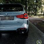 En BMW X3 xDrive30e hybrid SUV med karakteristiske baglygter parkerer på en våd vej midt i frodige grønne områder og viser sit sporty design og "m performance"-mærket, der antyder