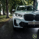 En sølvfarvet BMW X3 parkeret på en våd asfaltsti omkranset af græs og træer og fremviser dens elegante grill og karakteristiske forlygter med en frisk udendørs baggrund.