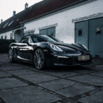 En elegant sort Porsche Boxster S med sin cabriolet-top nedad er parkeret på brosten, hvilket fremhæver dets sporty design på baggrund af en vintage hvid og blå bygning i skumringen.