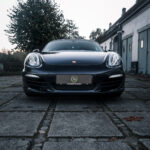 En elegant Porsche Boxster S-sportsvogn parkeret på en brostensbelagt sti, dens karakteristiske forlygter og emblem oplyst, med en baggrund af en tusmørkehimmel og historisk arkitektur.