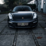 En slank sort Porsche Boxster S parkerer på en brostensbelagt indkørsel og viser sit ikoniske emblem og forlygter, der udstråler luksus i tusmørket.