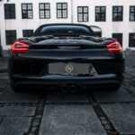 En slank sort Boxster S er parkeret på en brostensbelagt gårdhave og viser sit dynamiske bageste design på baggrund af en klassisk hvid bygning med sorte vinduesrammer, der inkarnerer en blanding af moderne teknik