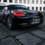 En slank sort Porsche Boxster S er parkeret på brostensbelagte fortov og viser dets sporty design og karakteristiske baglygter med baggrund af klassiske hvidtrimlede vinduer i en historisk bygning