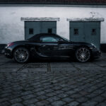 En elegant sort Porsche Boxster parkeret på en brostensbelagt overflade, i kontrast til den forsimplede baggrund af en gammel bygning med rustikke døre, under en overskyet himmel.