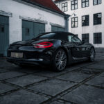 En slank sort Porsche 718 Boxster S parkeret i en gammel europæisk gårdhave, der praler med sit sporty design og luksuriøse tiltrækningskraft mod den kontrastfulde baggrund af historisk arkitektur.