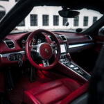 Et luksuriøst bilinteriør kan prale af en rig rød læderfinish med et slankt rat prydet med det karakteristiske Boxster S-badge, der antyder avanceret håndværk, sat op mod en historisk bygning