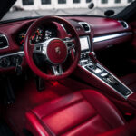 Luksuriøst rødt læderinteriør i en Porsche Boxster S-sportsvogn med en slank midterkonsol, moderne instrumentbræt og det ikoniske Porsche-emblem på rattet.