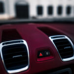 Et nærbillede af en Porsche Boxsters røde instrumentbræt, der fremhæver hazard light-knappen, ventilationsåbninger og et aux-kabel med en sløret bygningsfacade i baggrunden.