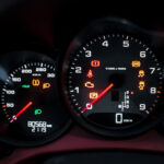 Oplyst Boxster S instrumentbræt, der viser speedometer, omdrejningstæller og forskellige advarselsindikatorer, med en sløret baggrund, der antyder, at det kan optages fra førerens perspektiv.