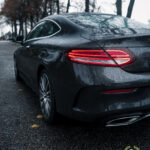 En slank sort Mercedes C300 Coupe parkeret på en våd vej, dens baglygter lyser rødt mod den humørfyldte baggrund af en overskyet dag, fremhævet af de glitrende dråber på dens
