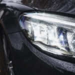 Et nærbillede af en slank, sort Mercedes C300's forlygte, der skinner med regndråber, og fremhæver avanceret lysteknologi midt i en våd, reflekterende overflade.