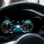 En moderne Mercedes C300 Coupes digitale instrumentbræt oplyst med et speedometer og brændstofmåler, set gennem en regndråbeindsmurt forrude, hvilket fremkalder en følelse af fokuseret kørsel i dårligt vejr.