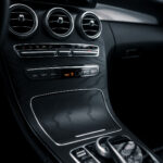 En luksusbils interiør, specifikt Mercedes C300 Coupe, viser en slank midterkonsol med en fremtrædende infotainmentskærm, cirkulære luftåbninger og en blank sort finish med metalliske accenter. Det