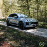En metallisk sølv Porsche Macan GTS parkeret på en grussti, omgivet af frodige grønne træer, der fremhæver kontrasten mellem luksus SUV-teknik og det naturlige miljø.
