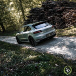 En Porsche Macan GTS er parkeret på en grussti i en solsprættet skov, og viser dets elegante design og terrænegenskaber med en bunke afhuggede træstammer i baggrunden, ev.