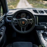 Førerens perspektiv af et Porsche Macan GTS-interiør med et læderrat, instrumentpanel og midterkonsol, placeret på en naturskøn vej midt i skovmiljøer i denne luksus-SUV.