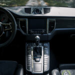 Indvendigt billede af Porsche Macan GTS med fokus på midterkonsollen og instrumentbrættet, der viser infotainmentsystemet, automatisk gearskifte og rat, med en vej synlig gennem