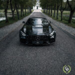 En elegant sort Mercedes AMG GT C Roadster parkeret på en fredfyldt allé med træer, der afspejler den overskyede himmel på dens polerede motorhjelm, der udstråler luksus og ydeevne.