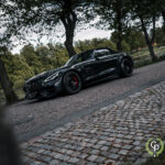 En elegant sort Mercedes-AMG GT Roadster parkeret på en brostensbelagt sti, der emmer af luksus og kraft, omgivet af fredfyldte parkgrønt under en overskyet himmel.