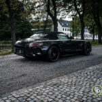 En slank sort Mercedes AMG GT cabriolet sportsvogn parkeret på en brostensbelagt sti, der praler med sit aggressive design og luksushjul, omgivet af rolig parklandskab med træer.