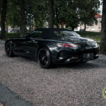 En elegant sort Mercedes AMG GT C Roadster parkeret på en brostensbelagt gade ved siden af en rolig vandvej med træer og en klassisk bygning i europæisk stil i baggrunden.