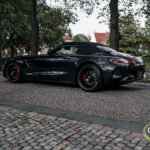 En elegant sort Mercedes AMG GT roadster med røde bremsekalipre parkeret på en brostensbelagt vej, omgivet af frodige grønne træer og traditionel europæisk arkitektur.