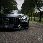 En elegant sort Mercedes Amg GT Roadster parkeret på en brostensbelagt sti og fremviser sin kraftfulde frontgrill og emblem under en baldakin af træer, der emmer af luksus og ydeevne.
