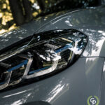 Et nærbillede af en luksus-SUV's slanke forlygte med indviklede designelementer, reflekterende solbeskinnede træer, antydning af præcisionsteknik og BMW X3 xDrive30e plug-in hybrid's høje