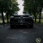 En sort Mercedes GLE350de SUV er parkeret på en asfaltvej, indrammet af frodige grønne træer, og viser dens imponerende gitter og karakteristiske Mercedes-emblem under en overskyet himmel.