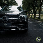 En skinnende sort Mercedes GLE350de SUV parkeret på en våd vejkant, dens krom-accenter afspejler det frodige grønne område under overskyet himmel.