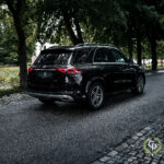 En slank sort Mercedes GLE350de parkeret på en brostensbelagt gade ved siden af en fredfyldt park og viser dens blanke finish og moderne design under en baldakin af frodige træer på en overskyet dag.