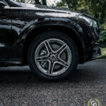 En luksusbil, specifikt Mercedes GLE350de hybrid SUV, med skinnende alufælge parkeret på en asfalteret vej, der viser sit slanke design og kontinentale dæk på baggrund af sløret grønt løv.