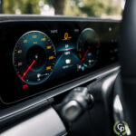 En moderne SUV's instrumentbræt, der fokuserer på det oplyste speedometer og omdrejningstæller på en Mercedes GLE350de hybrid, med udsigt til en trækantet gade udenfor, hvilket antyder en scene af ro
