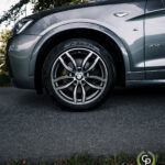 Et nærbillede af en BMW X3's forhjul, der viser dens stilfulde alufælge og Pirelli P Zero-dæk, delvist oplyst af naturligt lys på baggrund af udendørs.