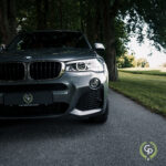 En sølvfarvet BMW X3 SUV med brugerdefineret præstationsmærke, der er parkeret på en træklædt vej, der viser sit slanke design og imponerende frontgitter mod en kulisse af frodige grønne områder.