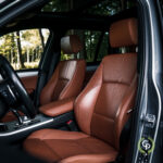 Luksus SUV-interiør med elegante solbrune lædersæder med fine syede detaljer, placeret ved siden af en åben dør midt i en fredfyldt skovkulisse, der viser BMW X3-modellen.