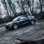 En elegant Model Y-elbil, muligvis med AWD, parkeret på en grynet betongrund med industriaffald i baggrunden under en humørfyldt, overskyet himmel.