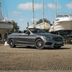 En stilfuld Mercedes C43 Cabriolet parkerer selvsikkert blandt forankrede yachts og inkarnerer luksus og essensen af fritid under en klar blå himmel.