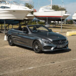 En slank, mørkegrå Mercedes C43 Cabriolet parkeret ved en lystbådehavn med en baggrund af både på tørdokker under en klar blå himmel, der viser luksus til lands og til vands.