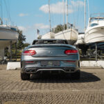 En sølvfarvet Mercedes C43 Cabriolet parkeret ved en lystbådehavn, med dets luksuriøse design sidestillet med en række forankrede sejlbåde under en klar blå himmel.