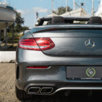 En Mercedes C43 Cabriolet med et bilpræstationsmærke parkeret på en indkørsel, omgivet af forankrede yachter, under en delvist overskyet himmel, der viser luksus og fritid.