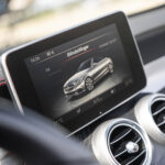 En moderne Mercedes C43 Cabriolets infotainmentsystem viser køretøjsindstillinger på dansk med slank grafik af bilen, sat mod en sløret baggrund af instrumentbrættets ventilationsåbninger og rat
