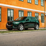 En elegant grøn Jaguar F-Pace, en luksus-SUV, parkeret på en brostensbelagt gade ved siden af en livlig orange bygning, der emmer af ydeevne og moderne design i historiske bymiljøer.