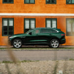En slank grøn Jaguar F-Pace, en luksus SUV, parkeret på en gade, med dens polerede overflade, der afspejler den orange facade af en vintage bygning, der legemliggør en blanding af moderne mobilitet og gammeldags