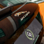Nærbillede af en klassisk Jaguar F-Pace's emblem på motorhjelmen, med en afspejling af arkitektoniske strukturer i den polerede brune lak, der viser køretøjets luksuriøse arv og omhyggelige vedligeholdelse.