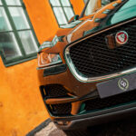 En Jaguar F-Pace SUV, i en karakteristisk brændt orange nuance, er parkeret i nærheden af en bygning med salviegrønne vinduer, der viser sin luksusgrill og elegante forlygter, der udstråler sofistikering og kraft.